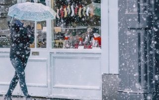 Frozen custard business planning time is best during the winter or fall months. This image shows a woman walking down a street in front of a shop window that is displaying stockings and gifts. It's snowing and she's holding an open umbrella to protect herself from the snowfall.
