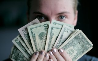 Frozen custard store profitability image. Woman is holding U.S. currency only her eyes and forehead appear behind the bills. The bills are fanned out and include several $1 bills, one $5 bill, two $20 bills and one $50 bill. Her fingernails have silver polish on them.