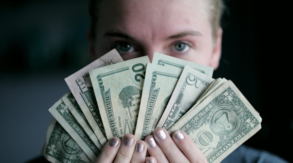 Frozen custard store profitability image. Woman is holding U.S. currency only her eyes and forehead appear behind the bills. The bills are fanned out and include several $1 bills, one $5 bill, two $20 bills and one $50 bill. Her fingernails have silver polish on them.