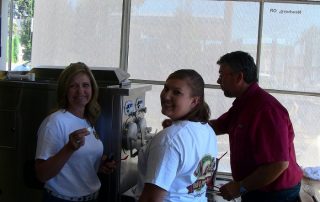 Gravity Fed Frozen Custard Machines Image: It's a man dressed in a red shirt training two women on how to operate a two barrel frozen custard machine. The women are wearing white t-shirts with the company logo, etc.