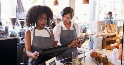 Ice cream shop new employee training shows two women in a food shop environment. One woman is showing the other how to operate the point of sale terminal. There's a tip jar on the counter.