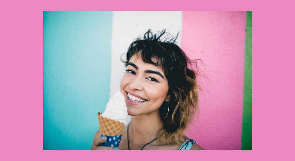 Maintaining frozen custard machines image. Young woman holding an ice cream cone. She is smiling and in front of a blue, white and pink background.