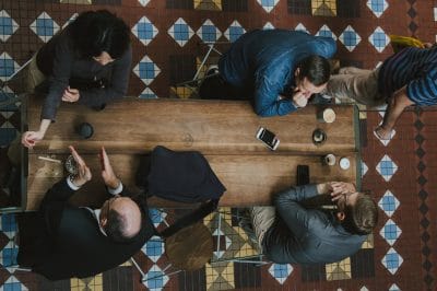 Meeting face to face image shows brown rectangular table. Three men and 1 woman is sitting at the table in discussion.